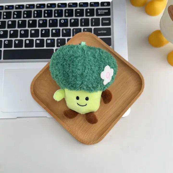 Broccoli plush keychain charm with flower detail and smiling face on a wooden tray